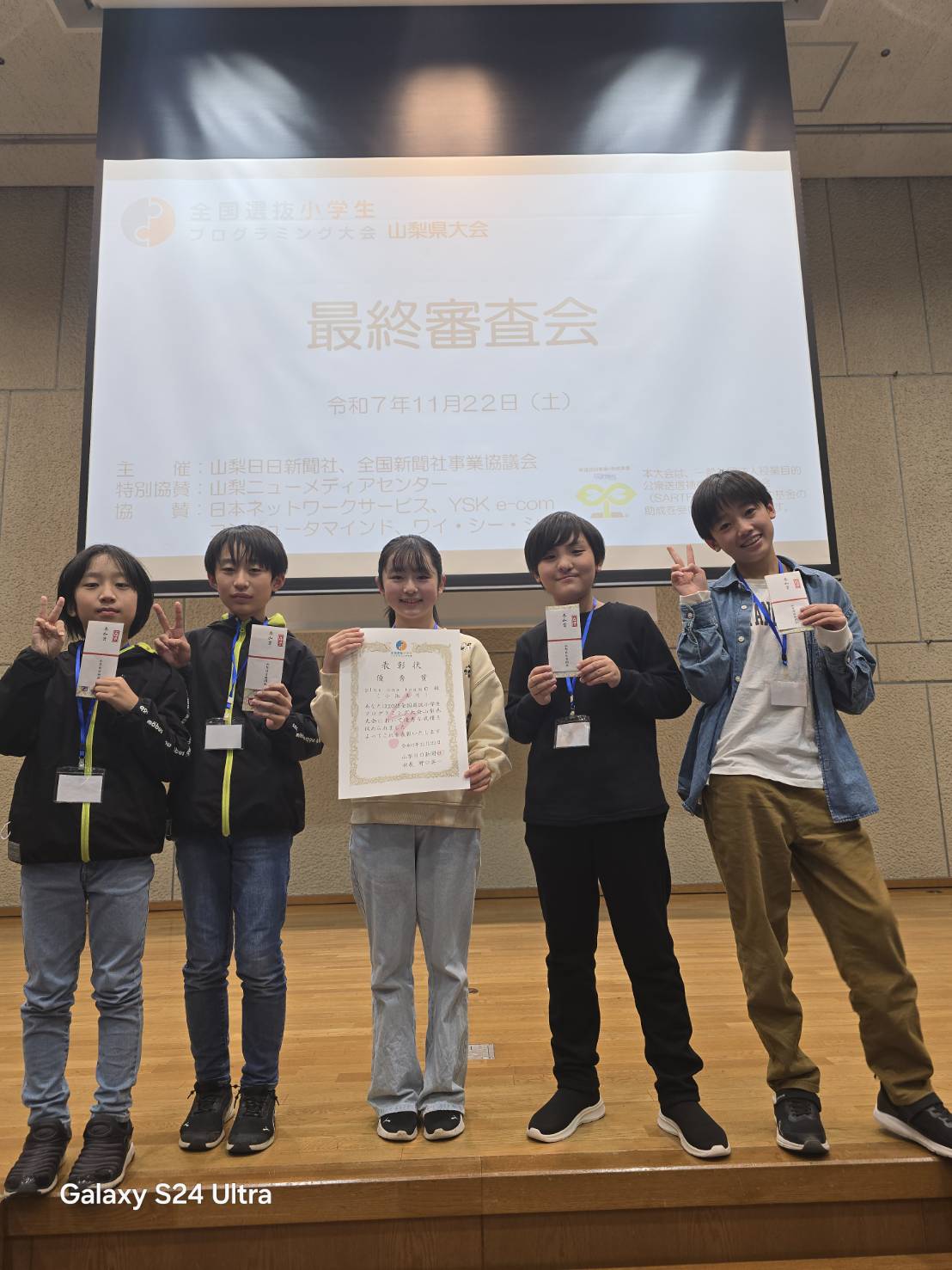 全国選抜小学生プログラミング大会 山梨県大会の最終審査会。ステージの前で、優秀賞の表彰状を持って並ぶ生徒たち5人